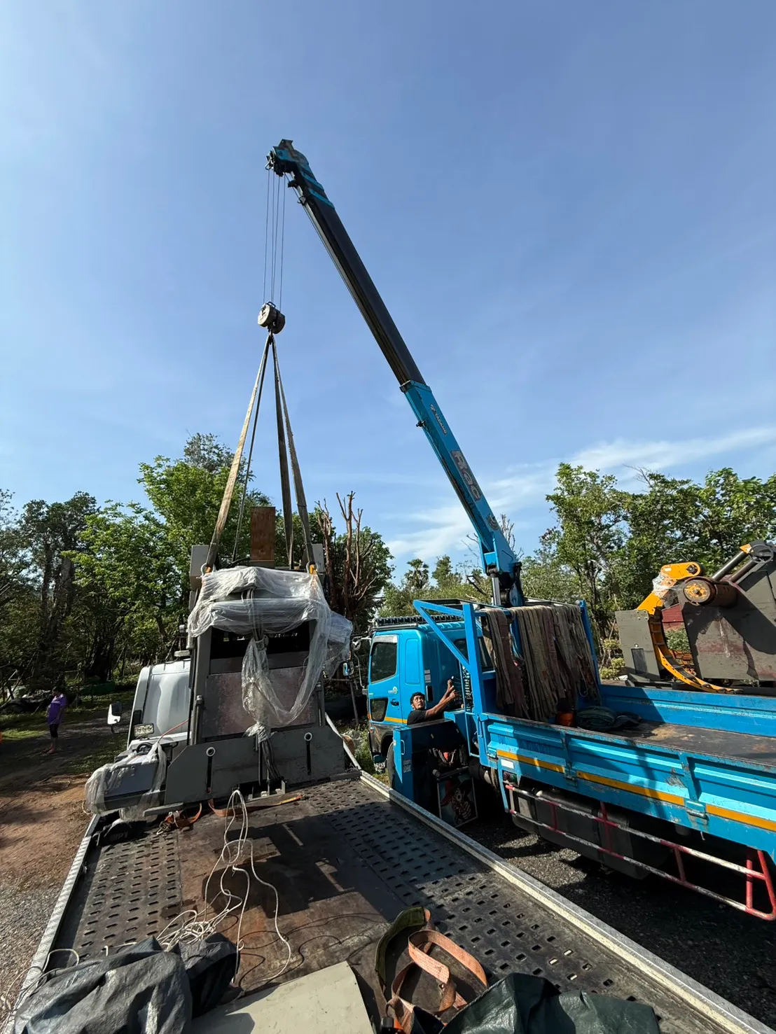 Blue crane mounted on a truck lifts a large wrapped industrial component onto a flatbed trailer outdoors